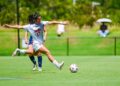 A female soccer player in a white uniform prepares to kick a ball on a sunny grass field at an outdoor stadium during a match.