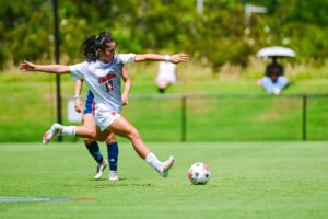 A female soccer player in a white uniform prepares to kick a ball on a sunny grass field at an outdoor stadium during a match.