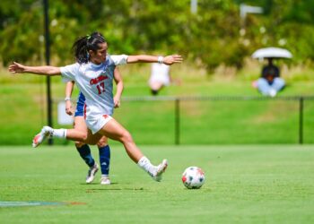 A female soccer player in a white uniform prepares to kick a ball on a sunny grass field at an outdoor stadium during a match.