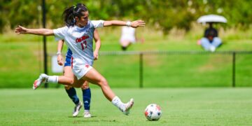 A female soccer player in a white uniform prepares to kick a ball on a sunny grass field at an outdoor stadium during a match.