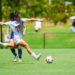 A female soccer player in a white uniform prepares to kick a ball on a sunny grass field at an outdoor stadium during a match.