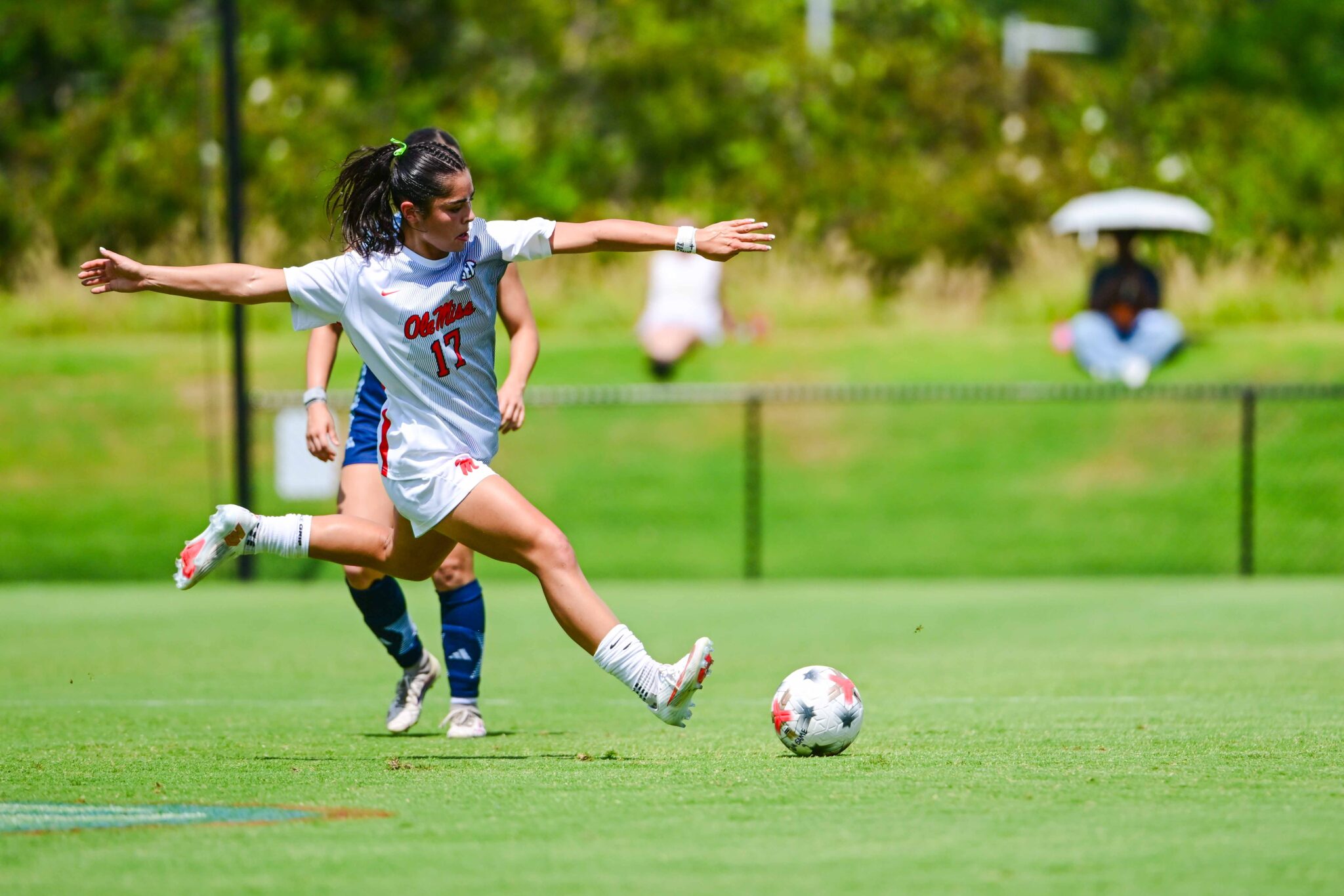 A female soccer player in a white uniform prepares to kick a ball on a sunny grass field at an outdoor stadium during a match.