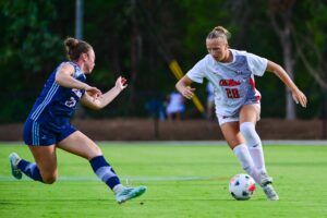A soccer player in a white uniform dribbles the ball across a green field at an outdoor stadium while an opposing athlete defends closely.