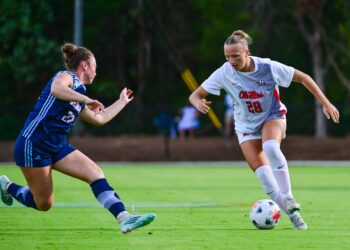 A soccer player in a white uniform dribbles the ball across a green field at an outdoor stadium while an opposing athlete defends closely.