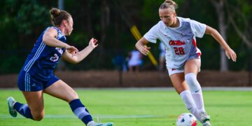 A soccer player in a white uniform dribbles the ball across a green field at an outdoor stadium while an opposing athlete defends closely.